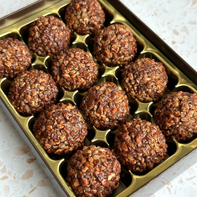 May include: A box of chocolate-covered treats with a crunchy texture. The treats are arranged in a grid pattern in a gold-coloured box with a brown lid.