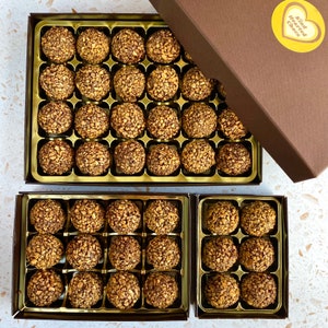 May include: Three boxes of chocolate truffles, each box containing a grid of individually wrapped truffles. The boxes are gold with a brown border and the top box has a yellow sticker with the text "Kind Hearted Chocs".