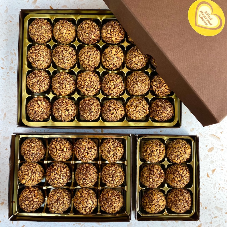 May include: Three boxes of chocolate truffles, each box containing a grid of individually wrapped truffles. The boxes are gold with a brown border and the top box has a yellow sticker with the text "Kind Hearted Chocs".