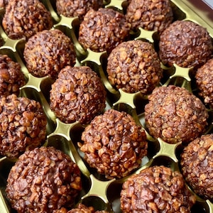 May include: A box of chocolate-covered treats with a crunchy texture. The treats are arranged in a grid pattern and are all the same size and shape. The chocolate is a dark brown colour and the crunchy texture is visible through the chocolate.