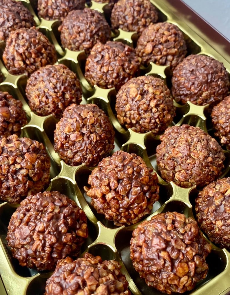 May include: A box of chocolate-covered treats with a crunchy texture. The treats are arranged in a grid pattern and are all the same size and shape. The chocolate is a dark brown colour and the crunchy texture is visible through the chocolate.