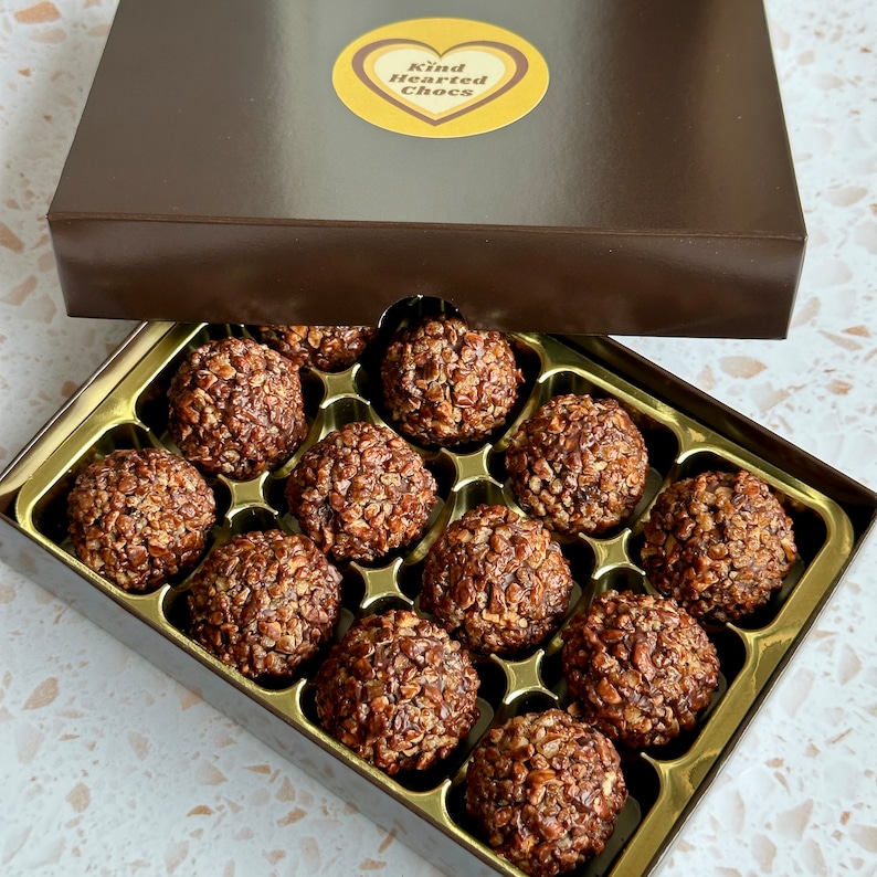 May include: A box of chocolate truffles with a gold-coloured tray. The truffles are covered in chocolate and have a crunchy texture. The box has a label that says "Kind Hearted Chocs".