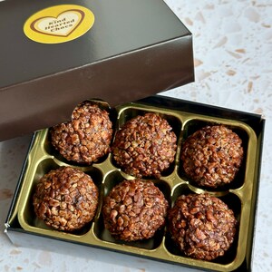 May include: A box of six chocolate truffles with a brown lid and a gold heart sticker that says "Kind Hearted Chocs". The truffles are covered in chocolate and have a crunchy texture.