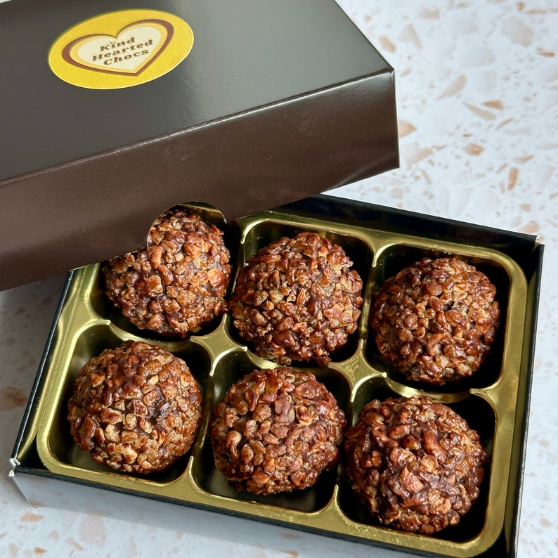 May include: A box of six chocolate truffles with a brown lid and a gold heart sticker that says "Kind Hearted Chocs". The truffles are covered in chocolate and have a crunchy texture.
