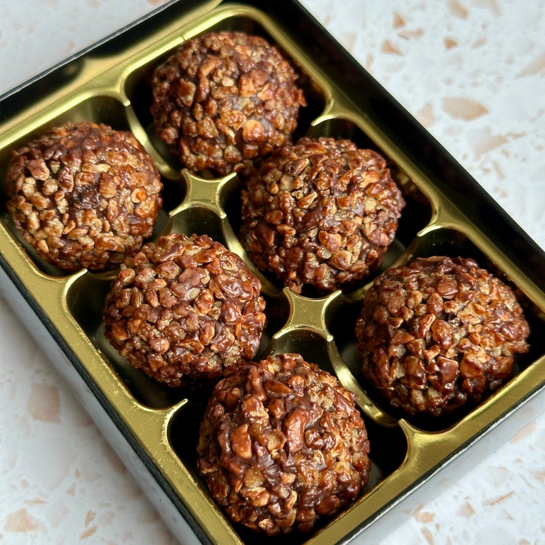 May include: Six chocolate-covered cereal treats in a gold and black box. The treats are round and have a rough texture.