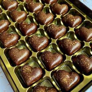 May include: A box of twenty-four heart-shaped dark chocolates arranged in a gold tray. Each chocolate has a smooth, glossy finish and is decorated with a fine, speckled gold dust pattern across the surface, creating a uniform appearance.