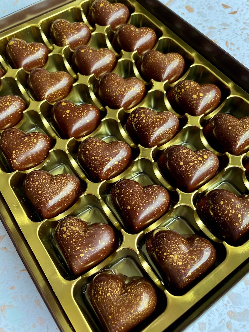 May include: A box of twenty-four heart-shaped dark chocolates arranged in a gold tray. Each chocolate has a smooth, glossy finish and is decorated with a fine, speckled gold dust pattern across the surface, creating a uniform appearance.