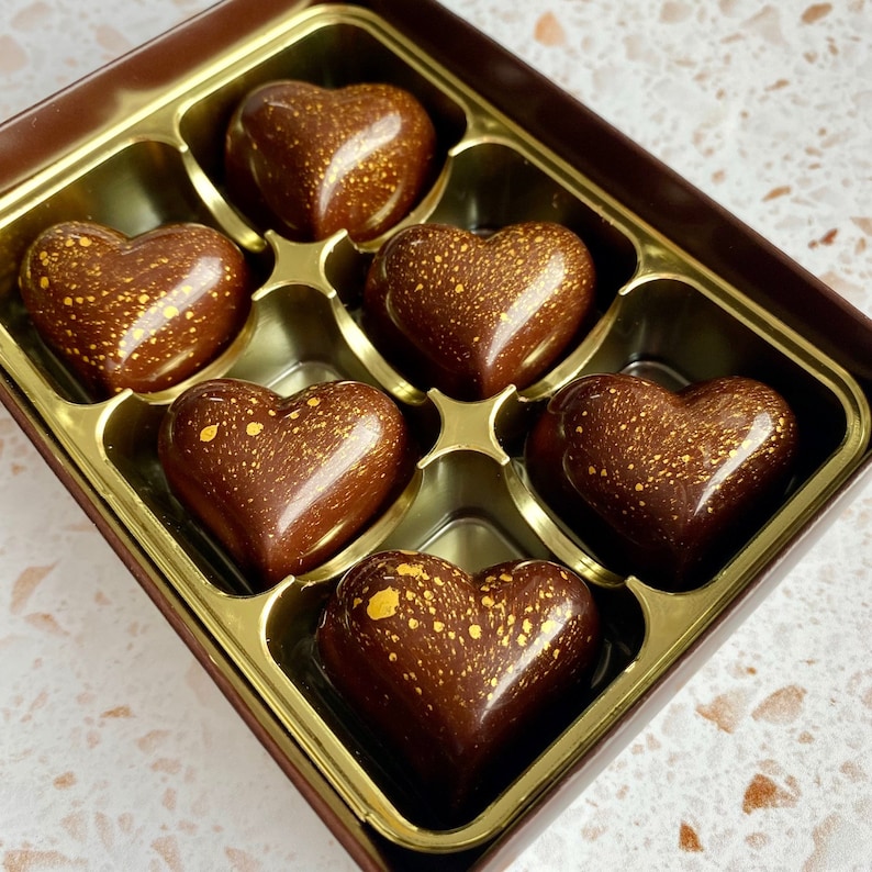 May include: A dark brown rectangular box containing six heart-shaped chocolates. Each chocolate has a smooth, glossy finish with a speckled gold metallic pattern across the surface. The chocolates are arranged in a gold-colored plastic tray.