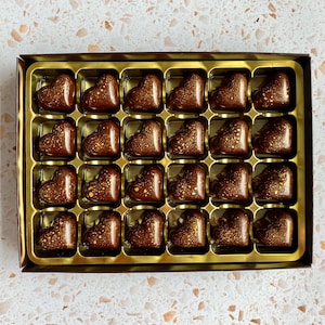 May include: A rectangular box containing twenty-four heart-shaped dark chocolate candies arranged in a gold plastic tray. Each chocolate features a smooth, glossy finish with decorative gold-colored speckles scattered across the surface.