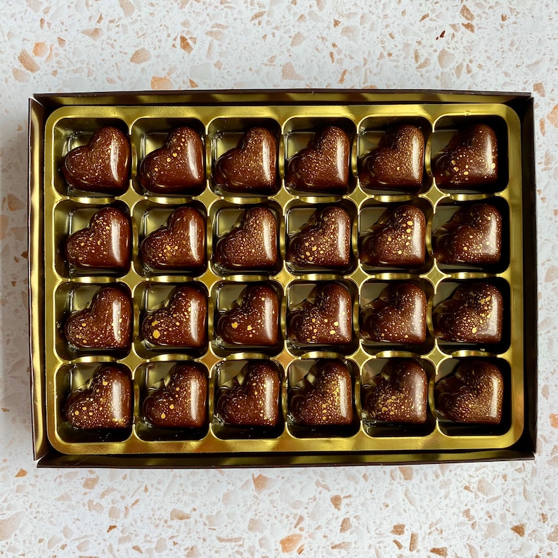 May include: A rectangular box containing twenty-four heart-shaped dark chocolate candies arranged in a gold plastic tray. Each chocolate features a smooth, glossy finish with decorative gold-colored speckles scattered across the surface.