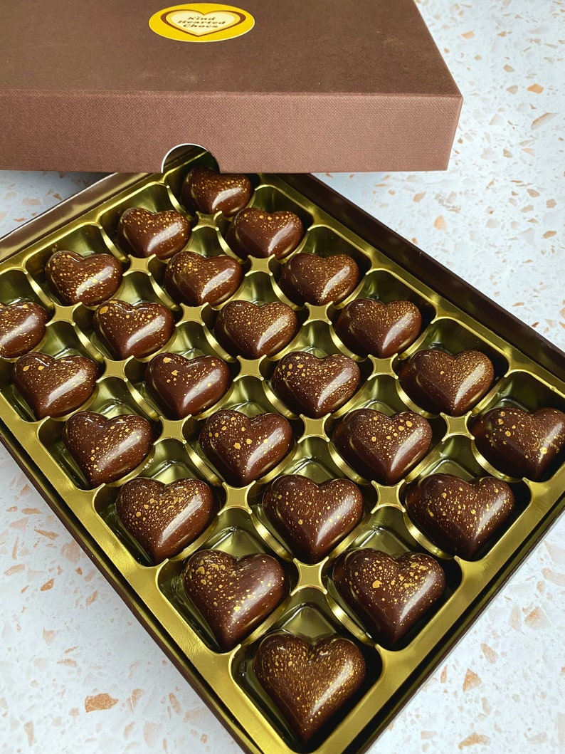 May include: A box of dark chocolate hearts with gold-colored speckles arranged in a gold-colored plastic tray. The brown box lid features a yellow heart-shaped sticker with the text Kind Hearted Chocs printed in black, partially covering the chocolates.