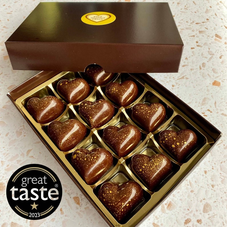 May include: A rectangular dark brown box containing twelve heart-shaped chocolates with gold-colored speckles. The lid features a yellow heart-shaped sticker with the text Kind Hearted Choco. A black circular logo reads great taste 2023.