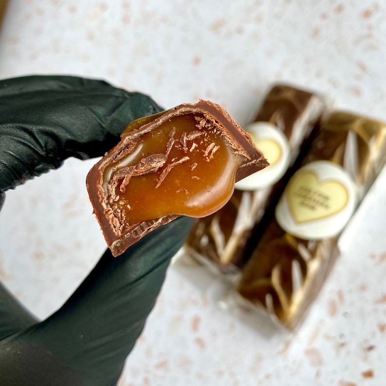 May include: A piece of milk chocolate candy broken in half to reveal a thick, golden-brown caramel filling. The chocolate has a smooth, dark brown exterior. Two additional wrapped chocolate bars with heart-shaped labels are visible in the background.