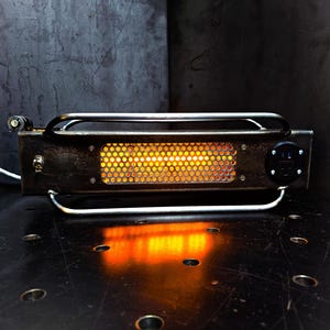 May include: A rectangular industrial-style lamp made of dark, weathered metal with a central perforated panel emitting a warm orange glow. The fixture features a silver metal handle frame, a toggle power switch, and a black electrical outlet port.