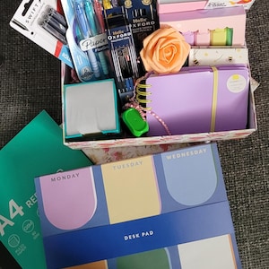 May include: A stationery gift box containing a variety of items including a weekly desk pad, a spiral-bound notebook, Helix Oxford ballpoint pens, Swift rollerball pens, a peach-coloured artificial rose, sticky notes, and a green highlighter.