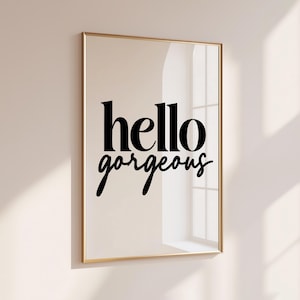 Peut inclure: Une affiche murale rectangulaire dans un cadre doré fin présentant le texte noir "hello gorgeous" dans une combinaison de polices serif et script. Le design est centré sur un fond beige clair, créant un affichage visuel propre et simple.