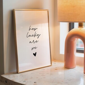 May include: A framed print with the words "how lucky are we" in black script, with a small heart below. The frame is gold, and the print is displayed on a speckled surface next to a pink lamp.