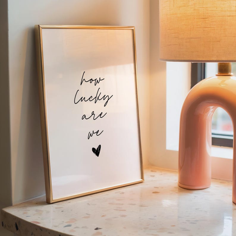 May include: A framed print with the words "how lucky are we" in black script, with a small heart below. The frame is gold, and the print is displayed on a speckled surface next to a pink lamp.