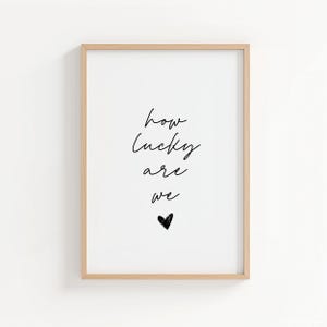 May include: A framed print with the words "how lucky are we" in black script, with a small black heart below. The frame is a light wood color, and the background is white.