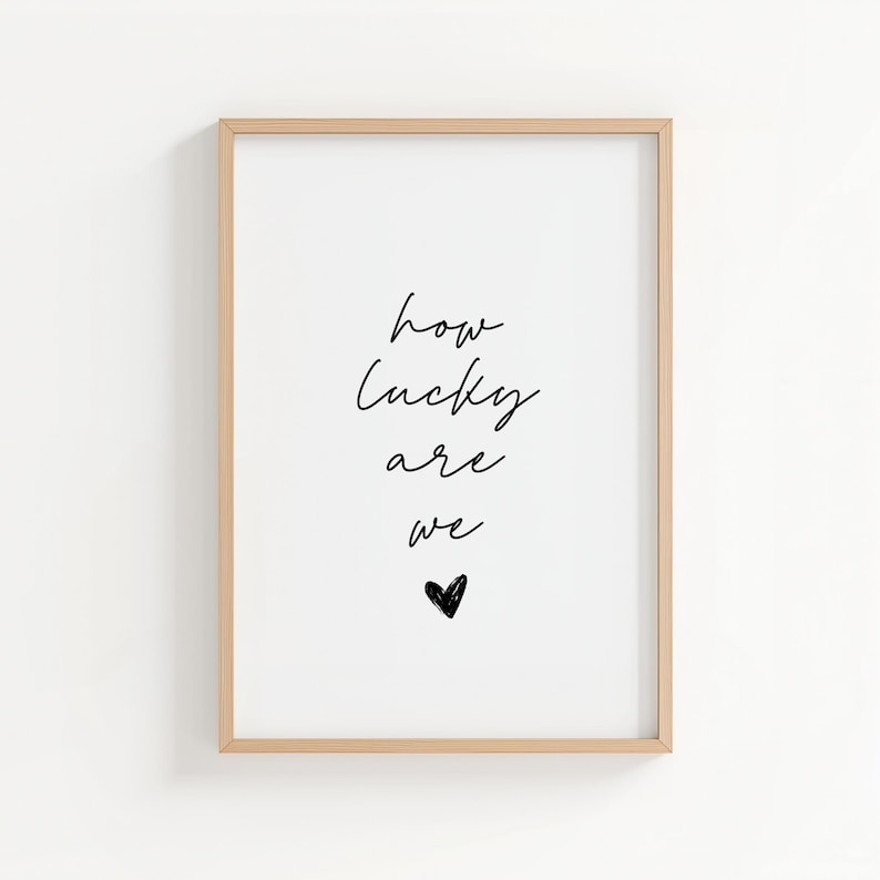 May include: A framed print with the words "how lucky are we" in black script, with a small black heart below. The frame is a light wood color, and the background is white.