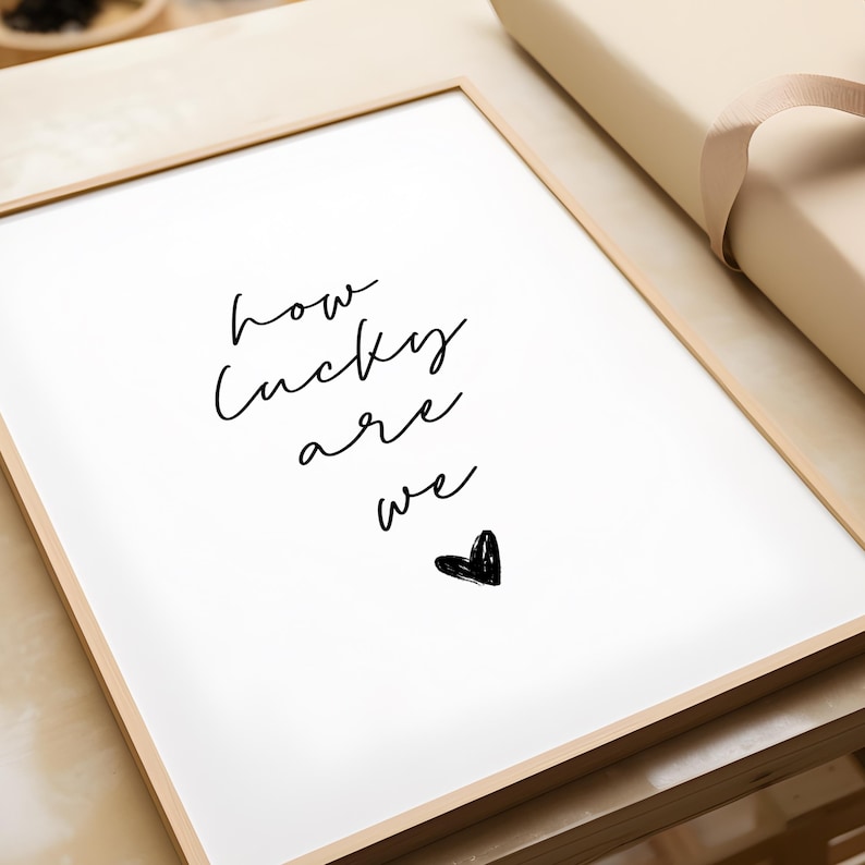 May include: A framed print with the words "how lucky are we" in black script. A small black heart is below the text. The frame is a light wood color, and the background is white.