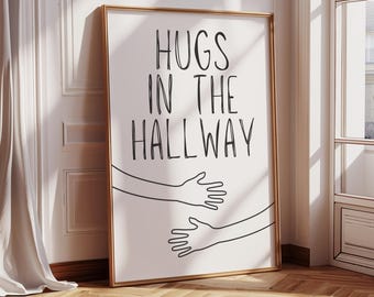 May include: A framed art print with the words "HUGS IN THE HALLWAY" in black lettering. Below the text are two line-drawn arms reaching out to embrace. The frame is gold, and the print is leaning against a white wall.
