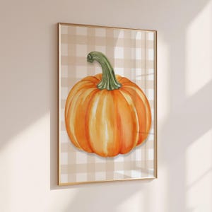 May include: A framed wall art print featuring a watercolour illustration of a bright orange pumpkin with a green stem, centred against a light beige and white checkered background. The artwork is enclosed in a thin, metallic gold frame.