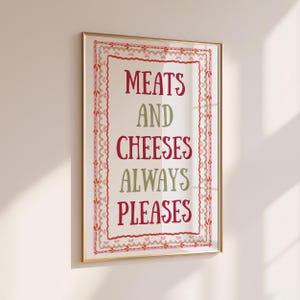 May include: A rectangular wall art print in a gold frame featuring the text MEATS AND CHEESES ALWAYS PLEASES in alternating red and olive green colours. The design is enclosed by a decorative border with small pink hearts and red stitching.