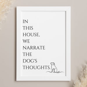 May include: White framed print with black text that reads "IN THIS HOUSE, WE NARRATE THE DOG'S THOUGHTS." A line drawing of a dog is in the bottom right corner. The background is a neutral beige.