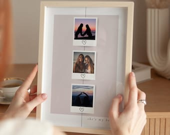 May include: A framed photo display with three Polaroid-style pictures. Each photo is attached with a string and features a heart drawn below. The bottom of the frame reads "she's my be". The frame is light wood.