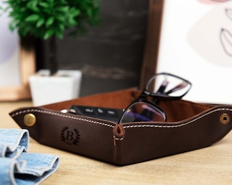 May include: A brown leather tray with a stitched edge and a brass rivet. The tray has a personalized initial "B" engraved in the center. The tray is filled with a pair of glasses and a key.