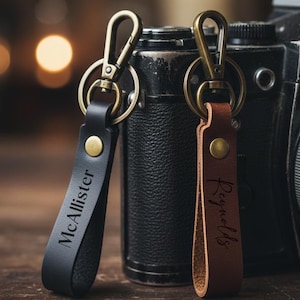 May include: Two leather keychains with metal clasps. One is black with "McAllister" engraved, the other is brown with "Reynolds" engraved. The keychains are displayed in front of a vintage camera.