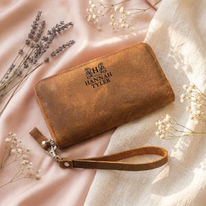 May include: A rectangular brown leather wallet with a wrist strap and a silver-toned metal zipper. The front is engraved with a decorative flourish, the letter H, and the name HANNAH TYLER in a dark, clear, sans-serif font.