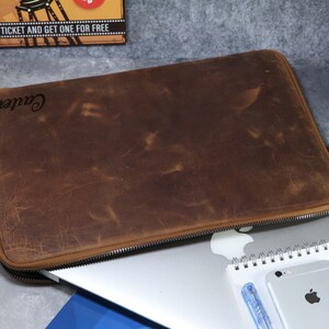 May include: A brown leather laptop sleeve with a distressed finish and the name Carter embossed in the upper left corner. The sleeve features a dark zipper closure with a leather pull tab, resting on top of a silver laptop, a notebook, and a smartphone.