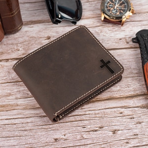 May include: A dark brown leather bi-fold wallet featuring a matte black cross emblem on the front corner. The wallet is constructed with visible light-colored stitching along the edges, providing a contrast to the smooth, distressed leather surface.