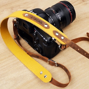 May include: A yellow leather camera strap with brown leather accents and a silver buckle. The strap is personalized with the name "JORDAN" in black lettering.