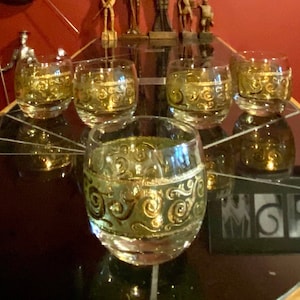 May include: Five gold-rimmed glasses with a swirling gold design on a black surface. The glasses are filled with a clear liquid. The background features a red wall with a framed picture and decorative swords.