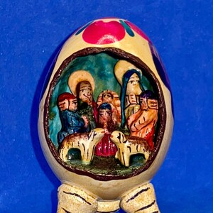 May include: A hand-painted ceramic egg-shaped ornament with a nativity scene inside. The egg is cream-colored with a floral design on top. The nativity scene features figures and two lambs. The ornament has three feet.