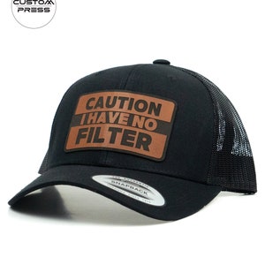 Puede incluir: Gorra de camionero negra con un parche de cuero marrón que dice "ATENCIÓN NO TENGO FILTRO".