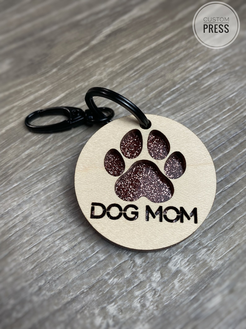 May include: A wooden keychain with a black metal ring and a paw print design. The paw print is filled with red glitter and the text "DOG MOM" is engraved below it.