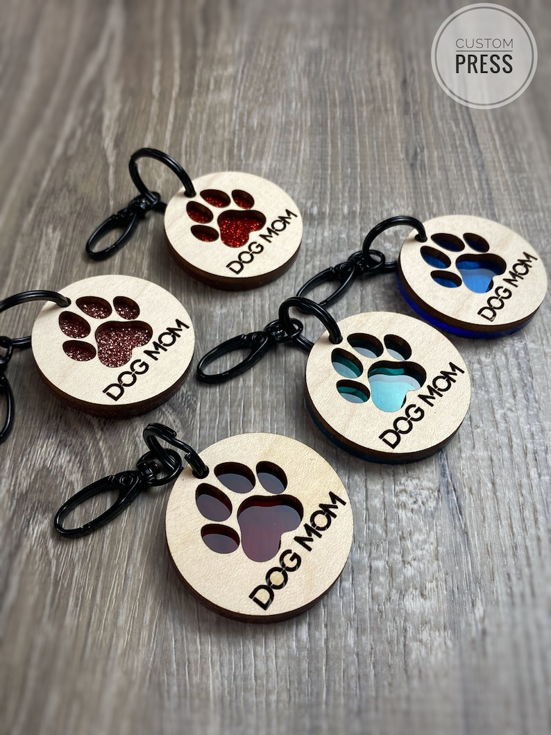 May include: Five wooden keychains with a paw print design. Each keychain has a different color resin insert in the paw print. The keychains say "Dog Mom" on them.
