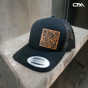 Puede incluir: Gorra de camionero negra y marrón con una parte trasera de malla negra. La gorra tiene un parche de cuero marrón con un código QR en blanco y negro. La gorra también tiene una pegatina plateada con el texto "The Authentic Yupoong Snapback Yupoong Since 1974".