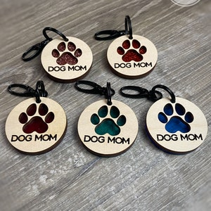 May include: Five wooden keychains with a paw print design. Each keychain has a different color resin insert in the paw print. The keychains say "DOG MOM" in black text.