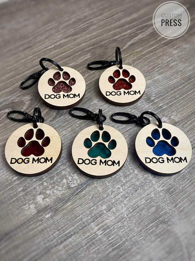 May include: Five wooden keychains with a paw print design. Each keychain has a different color resin insert in the paw print. The keychains say "DOG MOM" in black text.