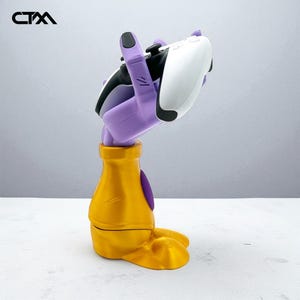 Può includere: Un supporto per controller di gioco dorato e viola a forma di mano che tiene un controller bianco e nero. Il supporto ha una base dorata e una mano viola con unghie nere. Il controller è bianco e nero.