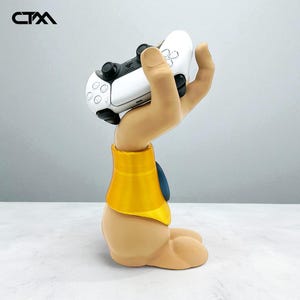 Puede incluir: Un mando de PlayStation 5 blanco y negro sostenido por un soporte beige con forma de mano y un cuello amarillo y azul. El soporte está sobre una superficie blanca. El logotipo "CTM" está en la esquina superior izquierda.