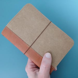 May include: A brown leather notebook with a tan cover and a brown elastic closure. The notebook is being held in a hand.