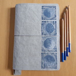 May include: A grey leather journal with a blue moon design on the cover. Four wooden pencils with blue tips are on the right side of the image.