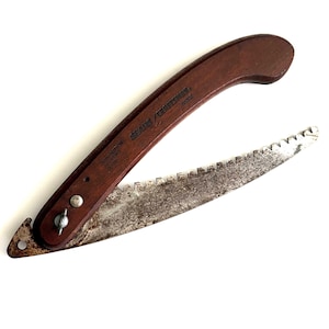 May include: A vintage Sears Craftsman folding saw with a dark brown wooden handle and a rusty, serrated blade. The saw is open, displaying the blade's teeth. The handle has the text "SEARS / CRAFTSMAN" and "PUSH BUTTON TO OPEN & CLOSE".