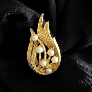 May include: A gold-toned brooch with a stylized flame design, adorned with six faux pearls. The brooch has a textured surface and is set against a black background, creating a striking contrast.